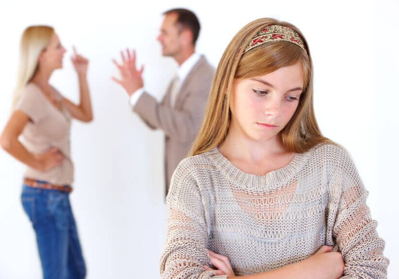 An unhappy young girl standing with her arms folded with her parents arguing in the background