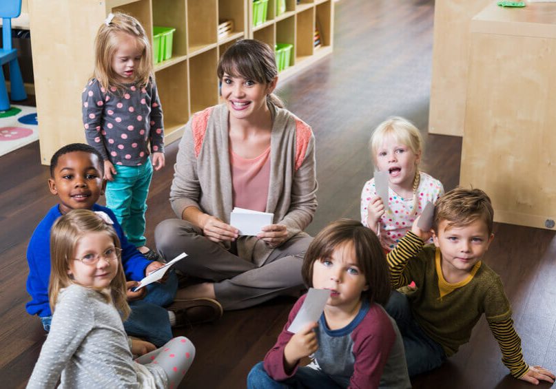 A preschool teacher sitting on the floor with a group of multi-ethnic children in a circle, smiling at the camera.  A little girl, a special needs child with down syndrome, is standing beside her wearing a gray shirt with pink polka dots.